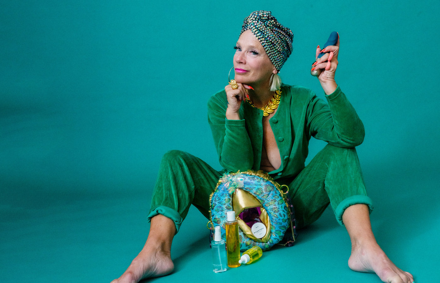 Lou on a green background in a green jumpsuit, with a vulva purse and holding the Deep Sea Blue STRONIC PETITE by Fun Factory